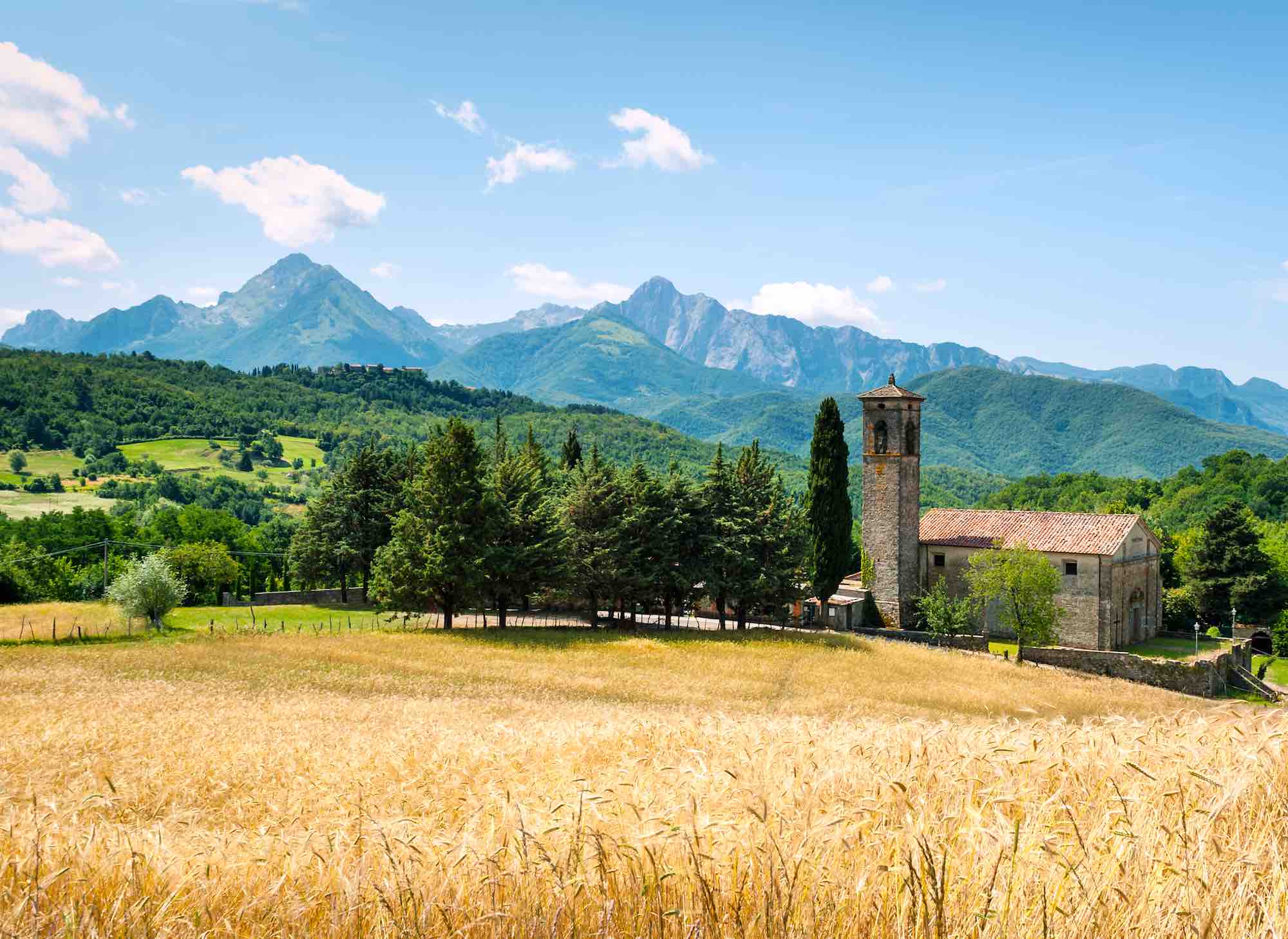 Chiesa in Lunigiana con campi di grano in primo piano e Alpi Apuane sullo sfondo