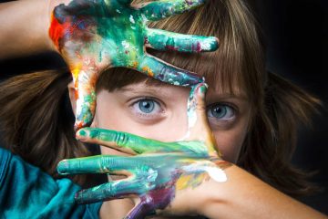 Bambina con occhi azzurri guarda attraverso le sue mani colorate con le tempere