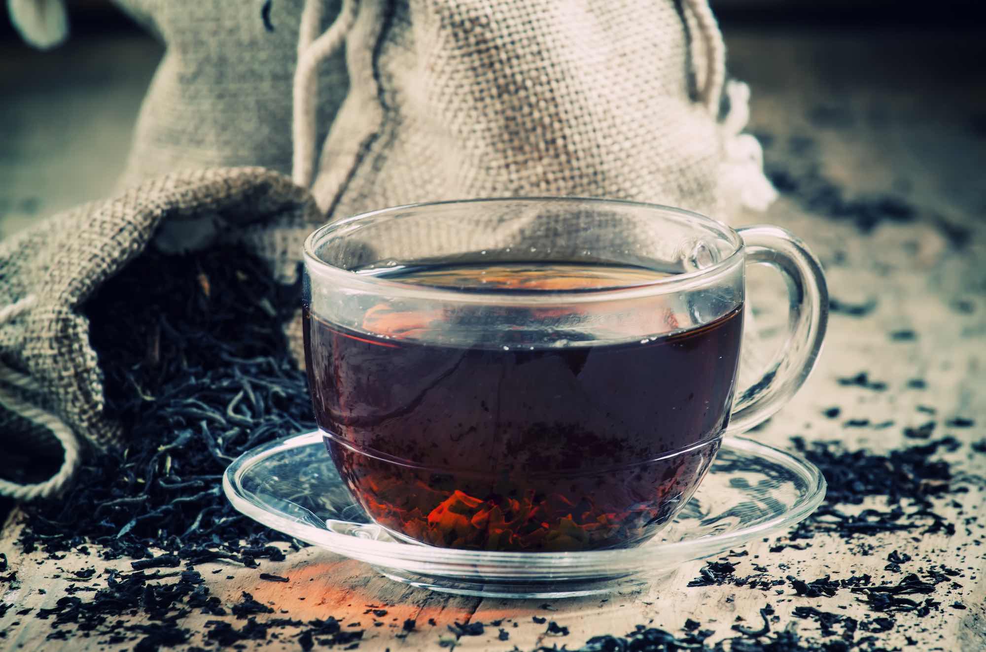 Tazza di vetro trasparente di tè nero e tè secco in bustine sullo sfondo di foglie di tè sparse sul vecchio sfondo di legno, messa a fuoco selettiva
