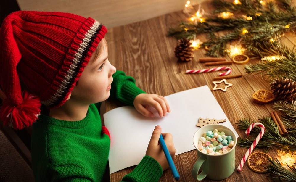 Bambino natalizio che scrive una lettera a Babbo Natale su un tavolo di legno marrone decorato con luci natalizie e abeti.
