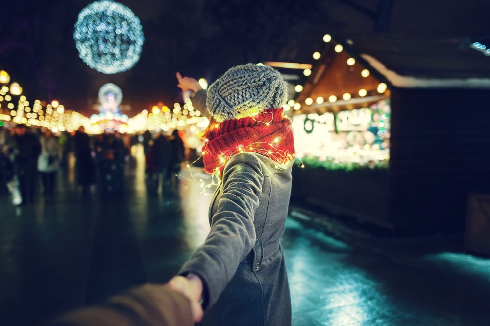 Giovane ragazza con cappotto invernale grigio, cappello e sciarpa rossa che tiene per mano il suo ragazzo e va alla fiera di Natale.
