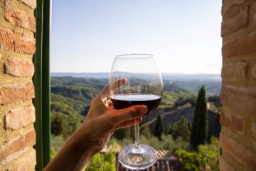 Donna che tiene in mano un bicchiere di vino rosso con un bellissimo paesaggio italiano sullo sfondo in una giornata di sole. Vista dalla finestra.
